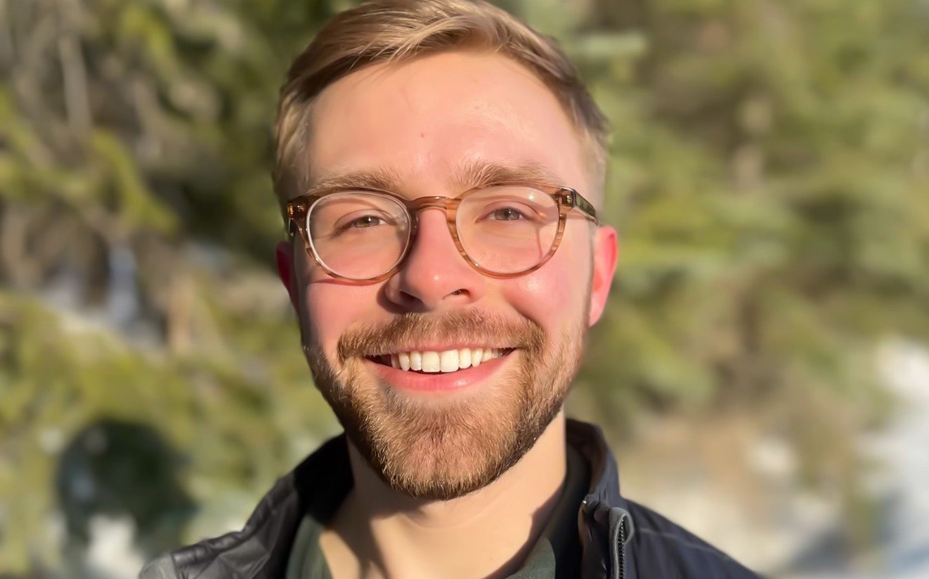 Man with short blonde hair, glasses and a goatee wearing a green button up shirt and dark jacket smiling in front of an outdoor nature scene