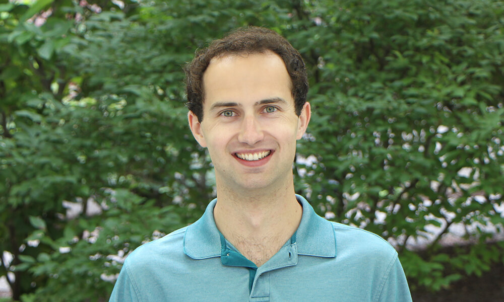 Man with short brown hair wearing a blue shirt stands in front of an outdoor background