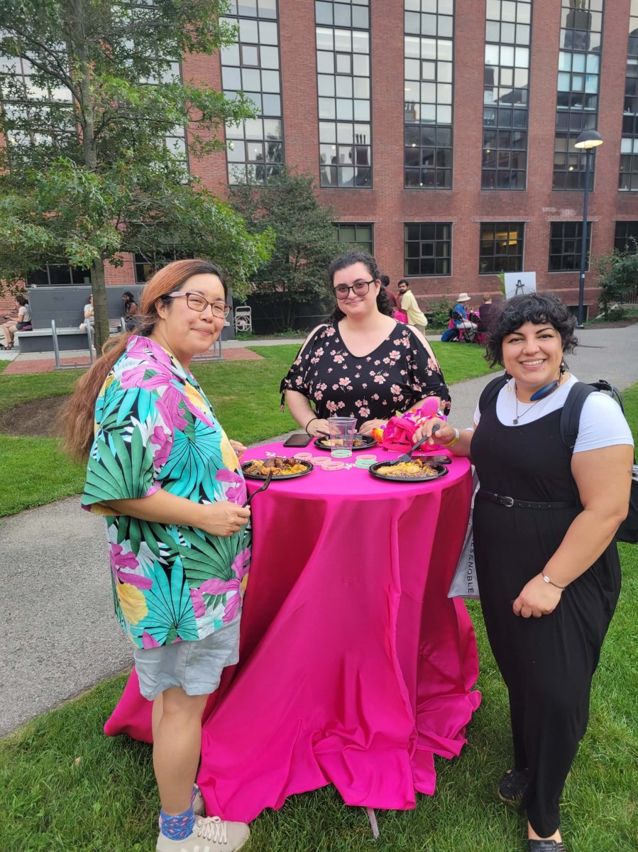 Three people standing around a pink table with a brick building in the background
