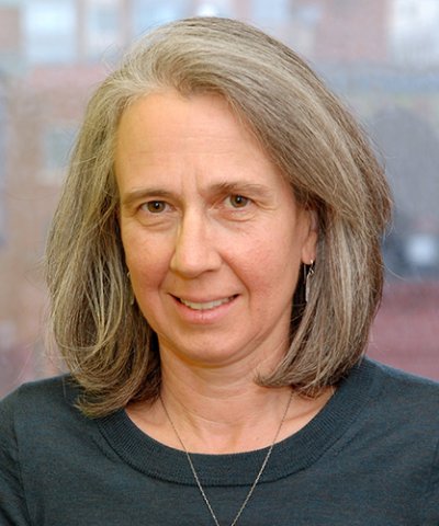 Person with shoulder length hair wearing a dark green blue shirt and a gold necklace